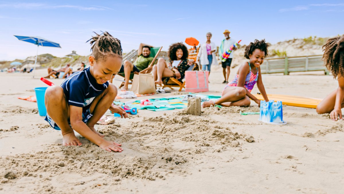 A group of children plays in the sand, building castles and laughing, while adults relax in the background on beach chairs under colorful umbrellas. The sunny beach scene is filled with joy, with rolling dunes and a clear blue sky completing the vibrant setting.